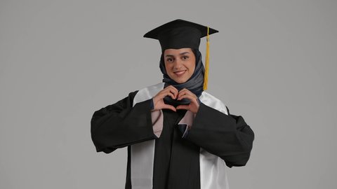 الشعور بالفخر والثقة بالنفس ، إنهاء الدراسة الجامعية، تحقيق حلم النجاح, الاحتفال بالتخرج من الجامعة، مفهوم الاجتهاد والتفوق ، بورترية لطالبة عربية خليجية اماراتيه محجبة ترتدي روب التخرج ترفع يديها للاعلى على شكل قلب، خلفية رمادية