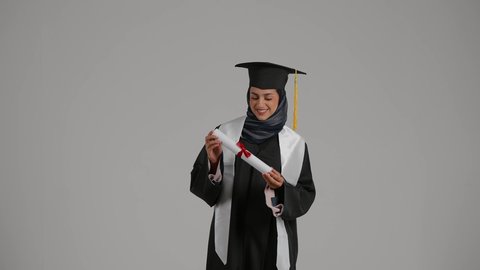 تحقيق حلم النجاح ، مفهوم الاجتهاد والتفوق، الشعور بالثقة بالنفس ، الاحتفال بإنهاء الدراسة الجامعية، بورترية لطالبة عربية خليجية اماراتيه محجبة ترتدي روب التخرج تقوم بامساك وثيقة النجاح بايماءات السرور، خلفية رمادية