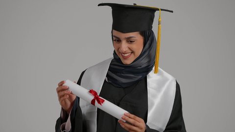 تحقيق حلم النجاح ، مفهوم الاجتهاد والتفوق، الاشاره الى وثيقه النجاح بتعابير الفخر، الشعور بالثقة بالنفس ، الاحتفال بإنهاء الدراسة الجامعية، بورترية مقرب لطالبة عربية خليجية اماراتيه محجبة ترتدي روب التخرج تقوم بامساك وثيقة النجاح بايماءات السرور، خلفية رمادية