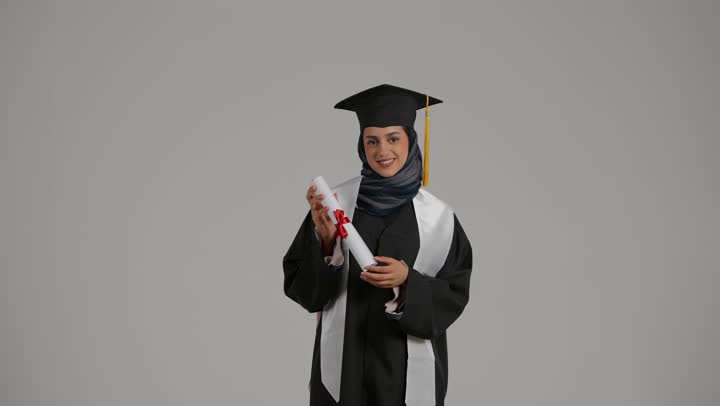 Achieving the dream of success, the concept of diligence and excellence, pointing to the success document with expressions of pride, feeling self-confidence, celebrating the completion of university studies, a portrait of a veiled Emirati Gulf Arab student wearing a graduation robe, raising the success document with gestures of pleasure, gray background
