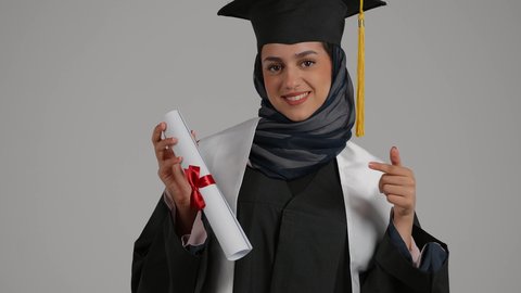 مفهوم الاجتهاد والتفوق، الاشاره الى وثيقه النجاح بتعابير الفخر ، تحقيق حلم النجاح, الشعور بالثقة بالنفس ، الاحتفال بإنهاء الدراسة الجامعية، بورترية مقرب لطالبة عربية خليجية اماراتيه محجبة ترتدي روب التخرج ترفع وثيقه النجاح بايماءات السرور