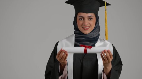 الشعور بالثقة بالنفس والفخر، الفرح بتحقيق النجاح، الاحتفال بإنهاء الدراسة الجامعية، مفهوم الاجتهاد والتفوق، بورترية مقرب لطالبة عربية خليجية اماراتيه محجبة ترتدي روب التخرج  ترفع وثيقه النجاح بايماءات السرور، خلفية رمادية