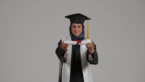 الفرح بتحقيق النجاح، الاحتفال بإنهاء الدراسة الجامعية، الشعور بالثقة بالنفس والفخر، مفهوم الاجتهاد والتفوق، بورترية لطالبة عربية خليجية اماراتيه محجبة ترتدي روب التخرج  ترفع وثيقه النجاح بايماءات السرور، خلفية رمادية