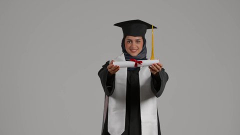 الاحتفال بإنهاء الدراسة الجامعية، الشعور بالثقة بالنفس والفخر، مفهوم الاجتهاد والتفوق، الفرح بتحقيق النجاح، بورترية لطالبة عربية خليجية اماراتيه محجبة ترتدي روب التخرج  ترفع وثيقه النجاح بايماءات السرور، خلفية رمادية