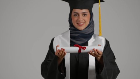الشعور بالثقة بالنفس والفخر، الاحتفال بإنهاء الدراسة الجامعية، مفهوم الاجتهاد والتفوق، الفرح بتحقيق النجاح، بورترية مقرب لطالبة عربية خليجية اماراتيه محجبة ترتدي روب التخرج  ترفع وثيقه النجاح بايماءات السرور، خلفية رمادية