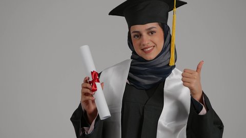 مفهوم الاجتهاد والتفوق، الشعور بالثقة بالنفس والفخر، الفرح بتحقيق النجاح، رفع قبضه اليد بايماءات السعاده ، بورترية مقرب لطالبة عربية خليجية اماراتيه محجبة ترتدي روب التخرج  ترفع وثيقه النجاح بايماءات السرور، خلفية رمادية