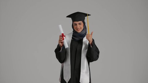 الشعور بالثقة بالنفس والفخر، الفرح بتحقيق النجاح، رفع قبضه اليد بايماءات السعاده ، مفهوم الاجتهاد والتفوق، بورترية لطالبة عربية خليجية اماراتيه محجبة ترتدي روب التخرج  ترفع وثيقه النجاح بايماءات السرور، خلفية رمادية
