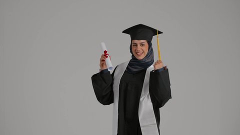 الفرح بتحقيق النجاح، رفع قبضه اليد بايماءات السعاده , الشعور بالثقة بالنفس والفخر، مفهوم الاجتهاد والتفوق، بورترية لطالبة عربية خليجية اماراتيه محجبة ترتدي روب التخرج  ترفع وثيقه النجاح بايماءات السرور، خلفية رمادية