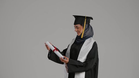 الشعور بالثقة بالنفس والفخر، مفهوم الاجتهاد والتفوق، الاحتفالات والمناسبات الجامعية، جودة التعليم في المملكة ، بورترية من الجانب لطالبة عربية خليجية اماراتيه محجبة ترتدي روب التخرج ترفع وثيقه النجاح بايماءات السعاده، خلفية رمادية