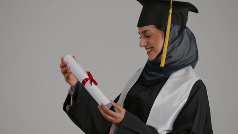 مفهوم الاجتهاد والتفوق، الاحتفالات والمناسبات الجامعية، الشعور بالثقة بالنفس والفخر، جودة التعليم في المملكة ، بورترية مقرب من الجانب لطالبة عربية خليجية اماراتيه محجبة ترتدي روب التخرج ترفع وثيقه النجاح بايماءات السعاده، خلفية رمادية