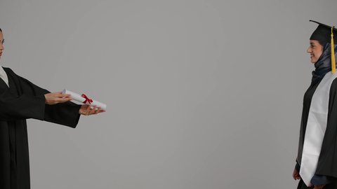 إنهاء الدراسة الجامعية، تحقيق حلم النجاح, الشعور بالفخر والثقه بالنفس ،مفهوم الاجتهاد والتفوق ، إيماءات الفرح والسرور، الاحتفال بالتخرج من الجامعة، بورترية من الجانب لطالبة عربية خليجية اماراتيه محجبة ترتدي روب التخرج تستلم وثيقه النجاح، خلفية رمادية