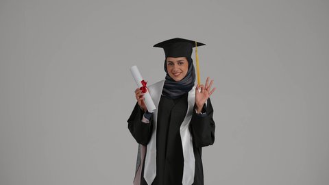 A historic moment in the lives of graduates, gestures of quality and excellence, university celebrations and events, expressions of pleasure and joy for achieving success, feeling of self-confidence and pride, the concept of diligence and excellence, a portrait of a veiled Emirati Gulf Arab student wearing a graduation robe, raising the success document with gestures of happiness, gray background