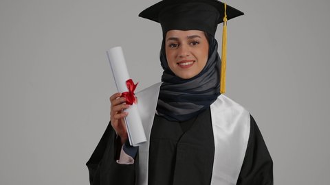 ايماءات الجودة والامتياز ،الاحتفالات والمناسبات الجامعية، الشعور بالثقة بالنفس والفخر، جودة التعليم في المملكة ، مفهوم الاجتهاد والتفوق، بورترية مقرب  لطالبة عربية خليجية اماراتيه محجبة ترتدي روب التخرج  ترفع وثيقه النجاح بايماءات السعاده، خلفية رمادية