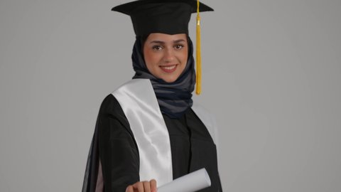 University celebrations and events, feeling of self-confidence and pride, quality of education in the Kingdom, concept of diligence and excellence, close-up portrait of a veiled Emirati Gulf Arab student wearing a graduation robe, raising the success document with gestures of happiness, gray background