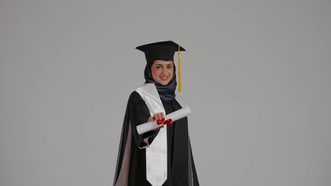 الاحتفالات والمناسبات الجامعية, تعابير السرور و الفرح لتحقيق النجاح، الشعور بالثقة بالنفس والفخر، جودة التعليم في المملكة ، مفهوم الاجتهاد والتفوق، بورترية لطالبة عربية خليجية اماراتيه محجبة ترتدي روب التخرج ترفع وثيقه النجاح بايماءات السعاده، خلفية رمادية