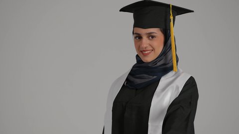 الشعور بالثقة بالنفس والفخر، تحقيق حلم النجاح ، جودة التعليم في المملكة ، الاحتفالات والمناسبات الجامعية, مفهوم الاجتهاد والتفوق، بورترية مقرب لطالبة عربية خليجية اماراتيه محجبة ترتدي روب التخرج  ترفع وثيقه النجاح بايماءات السعاده، خلفية رمادية