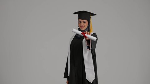 تحقيق حلم النجاح ، الشعور بالثقة بالنفس والفخر، جودة التعليم في المملكة ، الاحتفالات والمناسبات الجامعية, مفهوم الاجتهاد والتفوق، بورترية لطالبة عربية خليجية اماراتيه محجبة ترتدي روب التخرج  ترفع وثيقه النجاح بايماءات السعاده، خلفية رمادية