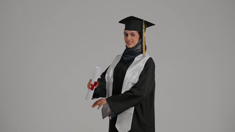 تحقيق حلم النجاح، الاحتفالات والمناسبات الجامعية، الشعور بالثقة بالنفس والفخر، جودة التعليم في المملكة ، مفهوم الاجتهاد والتفوق، بورترية لطالبة عربية خليجية اماراتيه محجبة ترتدي روب التخرج تقف بايدي متقاطعه، خلفية رمادية
