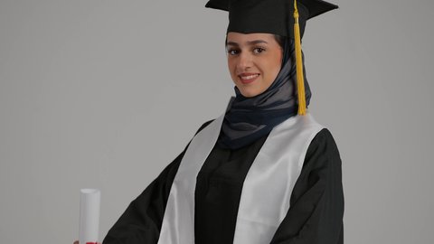 الاحتفالات والمناسبات الجامعية ، تحقيق حلم النجاح ، الشعور بالثقة بالنفس والفخر، جودة التعليم في المملكة ، مفهوم الاجتهاد والتفوق، بورترية مقرب لطالبة عربية خليجية اماراتيه محجبة ترتدي روب التخرج تقف بايدي متقاطعه، خلفية رمادية