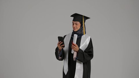 الاحتفالات والمناسبات الجامعية، توثيق اللحظات التذكاريه , تحقيق حلم النجاح ، الشعور بالثقة بالنفس والفخر، جودة التعليم في المملكة ، مفهوم الاجتهاد والتفوق،بورترية لطالبة عربية خليجية اماراتيه محجبة ترتدي روب التخرج تحمل بيدها وثيقه النجاح والهاتف المحمول، خلفية رمادية