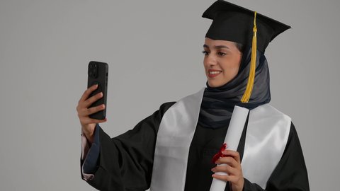 توثيق اللحظات التذكاريه ، الاحتفالات والمناسبات الجامعية ، الشعور بالثقة بالنفس والفخر، جودة التعليم في المملكة ، مفهوم الاجتهاد والتفوق،بورترية مقرب لطالبة عربية خليجية اماراتيه محجبة ترتدي روب التخرج ، تحمل بيدها وثيقه النجاح والهاتف المحمول، خلفية رمادية