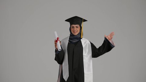 الاحتفال بتحقيق النجاح، مفهوم النجاح والتفوق، بورترية لطالبة عربية خليجية اماراتية ترتدي عباءه التخرج تحمل شهادة التخرج و تمسك بالقبعه بمشاعر الفرح والامتنان، الاحتفال بانهاء الدراسة الجامعية، خلفية رمادية