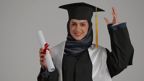 ايماءات البهجة والسعادة بالوصول لحلم التخرج، مفهوم النجاح والتفوق، بورترية مقرب لطالبة عربية خليجية اماراتية ترتدي عباءه التخرج تحمل شهادة التخرج و تمسك بالقبعه بمشاعر الفرح والامتنان، الاحتفال بانهاء الدراسة الجامعية، خلفية رمادية