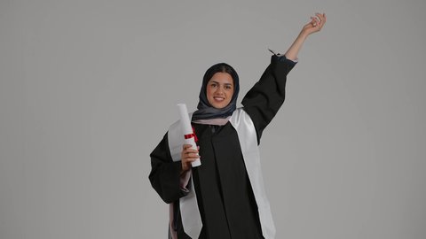 الاحتفال بإنهاء الدراسة الجامعية، مفهوم النجاح والتفوق، الشعور بالفخر والثقة بالنفس، بورترية لطالبة عربية خليجية اماراتية ترتدي عباءه التخرج تحمل شهادة التخرج و تقوم بالقاء قبعه التخرج الى الاعلي بمشاعر الفرح والامتنان، خلفية رمادية