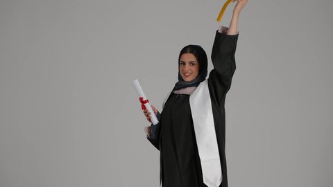 مفهوم النجاح والتفوق، الاحتفال بإنهاء الدراسة الجامعية، الشعور بالفخر والثقة بالنفس، بورترية لطالبة عربية خليجية اماراتية ترتدي عباءه التخرج تحمل شهادة التخرج و تقوم بالقاء قبعه التخرج الى الاعلي بمشاعر الفرح والامتنان، خلفية رمادية