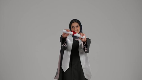 الاحتفال بالوصول إلى الأحلام وتحقيقها، بورتريه لخريجة عربية خليجية اماراتية محجبة مبتسمة ترتدي زي التخرج تقوم بامساك شهادة التخرج، شابة سعودية تحتفل بإنهاء الدراسة الجامعية، خلفية رمادية