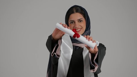 ايماءات البهجة والسعادة بالوصول لحلم التخرج، بورتريه مقرب لخريجة عربية خليجية اماراتية محجبة مبتسمة ترتدي زي التخرج تقوم بامساك شهادة التخرج، شابة سعودية تحتفل بإنهاء الدراسة الجامعية، ، خلفية رمادية