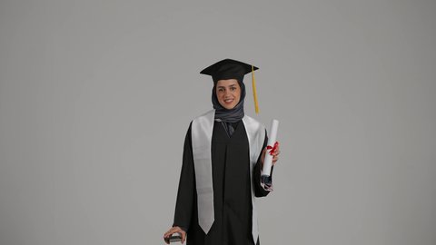 ايماءات الفرح بالتفوق، استخدام الأجهزة والتقنيات الحديثة, بورتريه لشابة عربية خليجية اماراتية ترتدي القبعة وعباءة التخرج بايماءات السعادة تمسك بيدها جوال و شهاده النجاح, بنت سعودية تقوم بعرض الهاتف المحمول بشاشة خضراء فارغة, خلفية رمادية