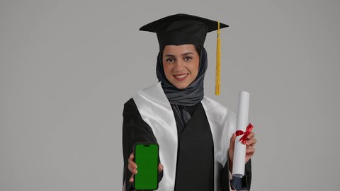 الشعور بالفخر والثقة بالنفس، استخدام الأجهزة والتقنيات الحديثة, بورتريه مقرب لشابة عربية خليجية اماراتية ترتدي القبعة وعباءة التخرج بايماءات السعادة تمسك بيدها جوال و شهاده النجاح, بنت سعودية تقوم بعرض الهاتف المحمول بشاشة خضراء فارغة, خلفية رمادية