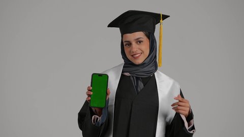 أهمية الدراسة الجامعية، استخدام الأجهزة والتقنيات الحديثة, بورتريه مقرب لشابة عربية خليجية اماراتية ترتدي القبعة وعباءة التخرج بايماءات السعادة تمسك بيدها الجوال و تشير اليه, بنت سعودية تقوم بعرض الهاتف المحمول بشاشة خضراء فارغة, خلفية رمادية