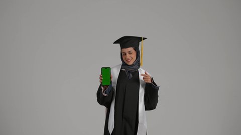 استخدام الأجهزة والتقنيات الحديثة, أهمية الدراسة الجامعية, بورتريه لشابة عربية خليجية اماراتية ترتدي القبعة وعباءة التخرج  بايماءات السعادة تمسك بيدهاهاتف محمول و تشير اليه, بنت سعودية تقوم بعرض الهاتف المحمول بشاشة خضراء فارغة, خلفية رمادية