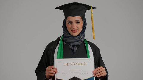الاحتفال بالتخرج من الفصل الدراسي, بورترية مقرب لطالبة عربيه خليجيه اماراتية ترتدي العباءه وقبعة التخرج تقوم بعرض شهادة النجاح  والنظر اليها بايماءات الفخر و السعادة، بنت سعودية تحتفل بإنهاء الدراسة الجامعية، الاهتمام بالأناقة والمظهر الخارجي، خلفية رمادية