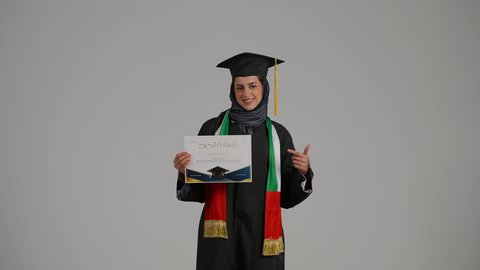مفهوم الاجتهاد والتفوق, بورترية لطالبة عربيه خليجيه اماراتية ترتدي العباءه وقبعة التخرج تقوم بالاشارة الى شهادة النجاح بايماءات الفخر و السعادة، بنت سعودية تحتفل بإنهاء الدراسة الجامعية، الاهتمام بالأناقة والمظهر الخارجي، خلفية رمادية