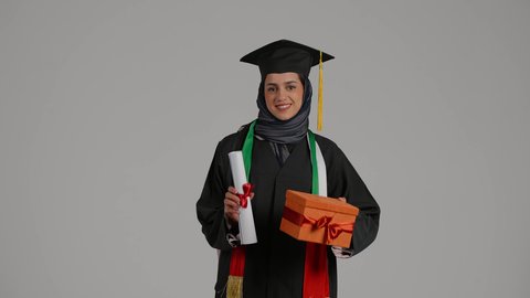 الاحتفال بإنهاء الدراسة الجامعية، بورتريه لخريجة عربية خليجية اماراتية محجبة ترتدي زي التخرج والقبعة تمسك بيدها وثيقة التخرج وعلبه هدايا تنظر إلى الكاميرا بتعابير السرور، شابة سعودية تحتفل بإنهاء الدراسة الجامعية، مناسبة واحتفال جامعي، خلفية رمادية