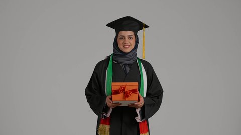 الشعور بالفرح والسعادة، الاحتفال بإنهاء الدراسة الجامعية،بورترية لطالبة عربية اماراتية خليجية ترتدي العباءه وقبعة التخرج ، بنت سعودية تحمل بيدها هدية تنظر إلى الكاميرا بإيماءات الرضا والسرور, مفهوم الاجتهاد والتفوق، خلفية رمادية