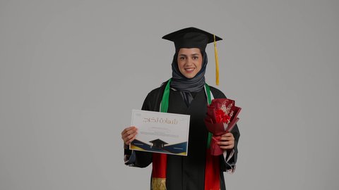 إيماءات السعادة بالوصول لحلم النجاح، بورتريه لخريجة عربية خليجية اماراتية محجبة ترتدي زي التخرج والقبعة تمسك بيدها وثيقة التخرج و بوكية ورد تنظر إلى الكاميرا بتعابير السرور، شابة سعودية تحتفل بإنهاء الدراسة الجامعية، مناسبة واحتفال جامعي، خلفية رمادية