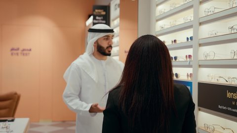 الاعجاب بالنظارات الشمسيه اثناء الشراء، متعة التسوق والشراء باختيار نظارة شمسيه مناسبة،  رجل عربي خليجي اماراتي يرتدي الكندوره يقوم بقياس نظاره ملونه، ارفف عرض النظارات، انواع وماركات النظارات الشمسيه، بائعه تقوم بمساعده الزبائن في محل النظارات
