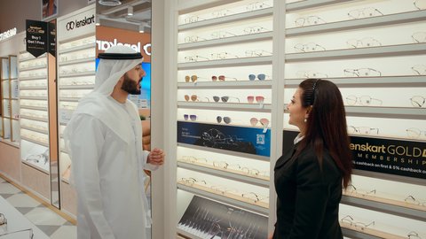 الاشاره الي النظارات الشمسيه، رجل اماراتي يرتدي الكندوره والغتره البيضاء يقوم بقياس النظارة الشمسيه المناسبة، الاهتمام بالمظهر الخارجي، التسوق وشراء النظارات الشمسيه من المولات، سيده اماراتيه تقوم بمساعده الزبائن في اختيار المقاسات المناسبه لهم
