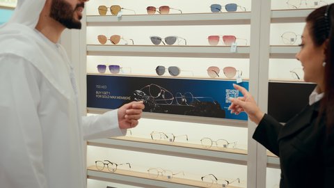 الاشاره الي النظارات الشمسيه، رجل اماراتي يرتدي الكندوره والغتره البيضاء يقوم بقياس النظارة الشمسيه المناسبة، الاهتمام بالمظهر الخارجي، التسوق وشراء النظارات الشمسيه من المولات، سيده اماراتيه تقوم بمساعده الزبائن في اختيار المقاسات المناسبه لهم