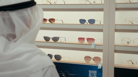 ارفف عرض النظارات الشمسيه الملونه، التسوق وشراء الاكسسورات في مولات الامارات، رجل عربي خليجي اماراتي يقوم بالاشاره الي نظاره ما بمساعده البائعه، ايماءات السرور الاعجاب