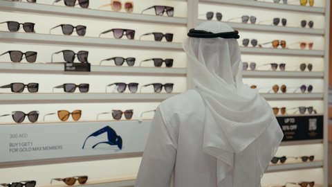 شاب اماراتي يرتدي الكندوره والغتره البيضاء يقوم بالنظر الي المرآه لقياس النظارة الشمسيه المناسبة، الاهتمام بالمظهر الخارجي، التسوق وشراء النظارات الشمسيه من المولات، عروض وتخفيضات، ايماءات الثقة بالنفس