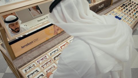 اختيار نظارة طبيه مناسبة، القيام بفحص النظارات الطبيه قبل الشراء، متعة التسويق والشراء باختيار نظارة طبيه مناسبة،  رجل عربي خليجي اماراتي يرتدي الكندوره يقوم بقياس نظاره طبيه، الاهتمام بالمنظر الخارجي، مفهوم ضعف النظر، خزانه عرض النظارات داخل المحل