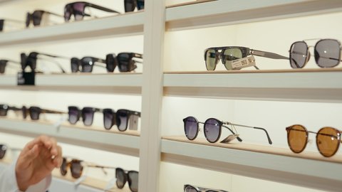 ارفف عرض النظارات داخل المحل، انواع وماركات النظارات الشمسيه العالميه، القيام ارتداء النظارات الشمسيه قبل الشراء، متعة التسويق والشراء باختيار نظارة شمسيه مناسبة،  رجل عربي خليجي اماراتي يرتدي الكندوره يقوم بقياس نظاره سوداء، الاهتمام بالمنظر الخارجي