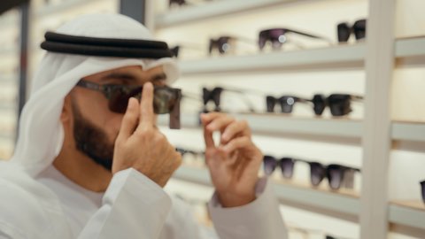 القيام ارتداء النظارات الشمسيه قبل الشراء، متعة التسويق والشراء باختيار نظارة شمسيه مناسبة،  رجل عربي خليجي اماراتي يرتدي الكندوره يقوم بقياس نظاره سوداء، ارفف عرض النظارات داخل المحل، انواع وماركات النظارات الشمسيه العالميه، ايماءه الثقه في النفس