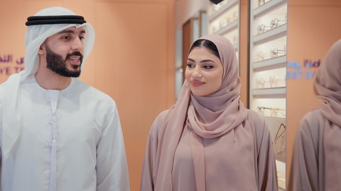 سيده اماراتيه ترتدي عباءه ورديه تستمتع باختيار وقياس نظارة طبيه مناسبة برفقه زوجها،  رجل عربي خليجي اماراتي يرتدي الكندوره يقوم باختيار نظاره طبيه لزوجته، ارفف عرض النظارات، انواع وماركات النظارات الطبيه، تحسين الرؤيه والنظر