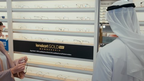 زوجان اماراتيان في متجر النظارات، سيده اماراتيه ترتدي عباءه ورديه تستمتع باختيار وقياس نظارة طبيه مناسبة برفقه زوجها،  رجل عربي خليجي اماراتي يرتدي الكندوره ونظاره طبيه يقوم باختيار نظاره طبيه، ارفف عرض النظارات، تحسين الرؤيه والنظر